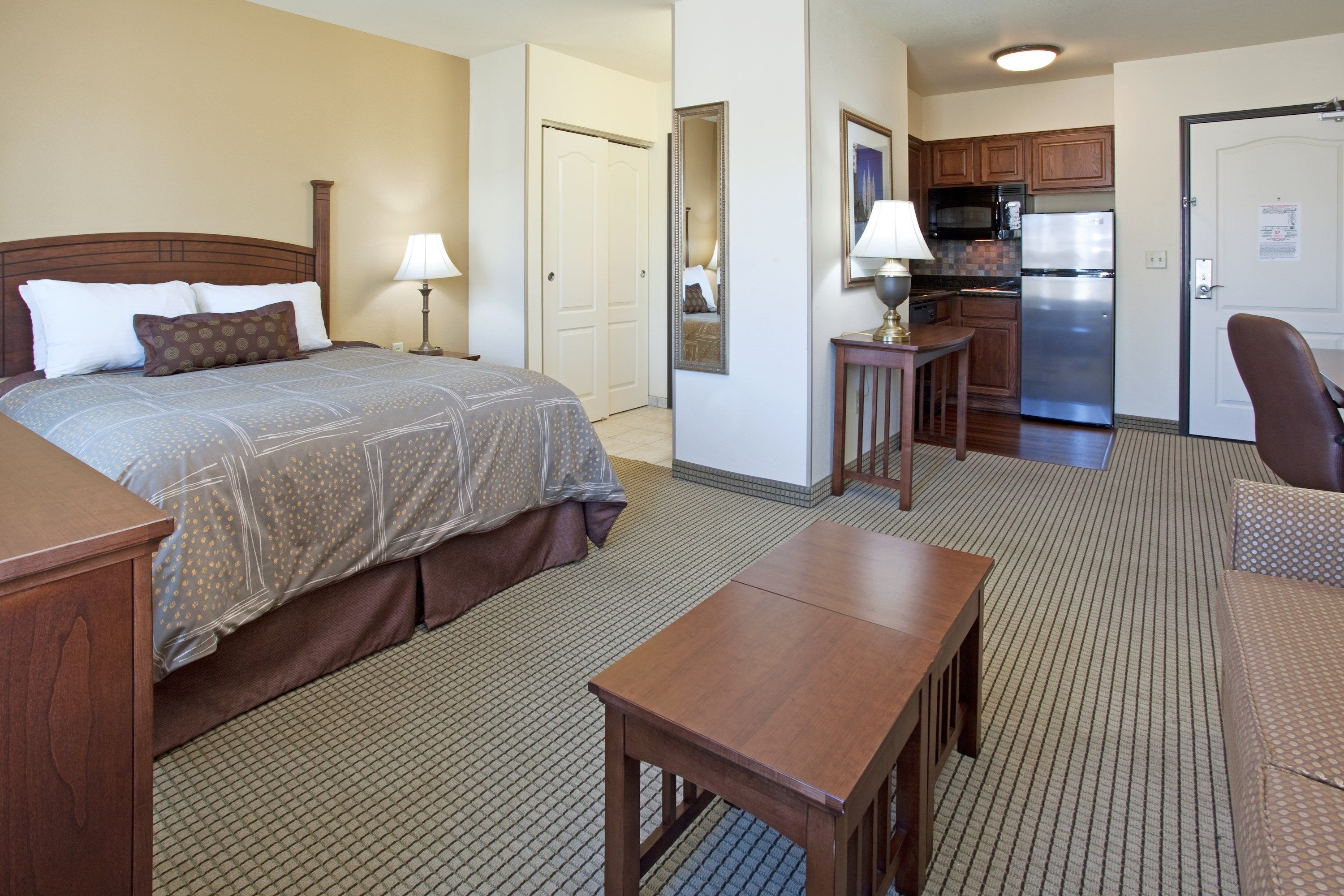 A hotel room with a bed, a desk, a chair, a small table, a lamp, a microwave, a refrigerator, and a kitchen area.