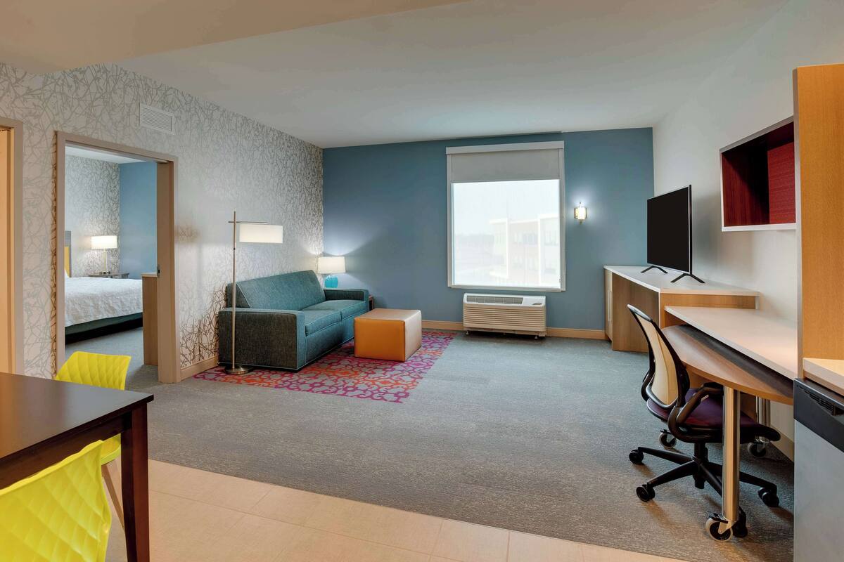 Quarto de hotel com cama, escrivaninha, cadeira, sofá, televisão e janela.
