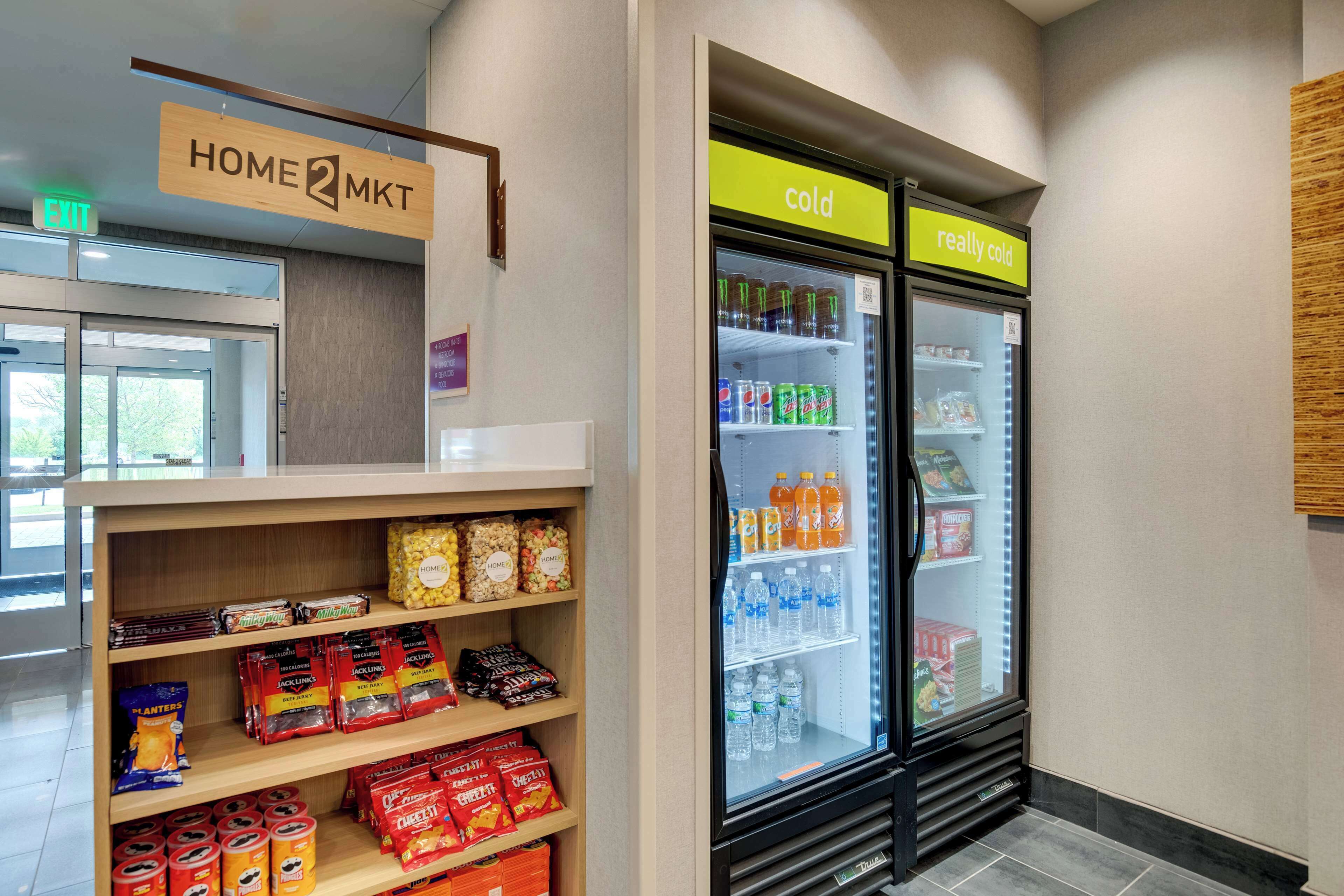 Un hall de hotel con un letrero 'Home 2 MKT', un refrigerador para bebidas 'frío' y 'muy frío', y estantes llenos de refrigerios y bebidas.