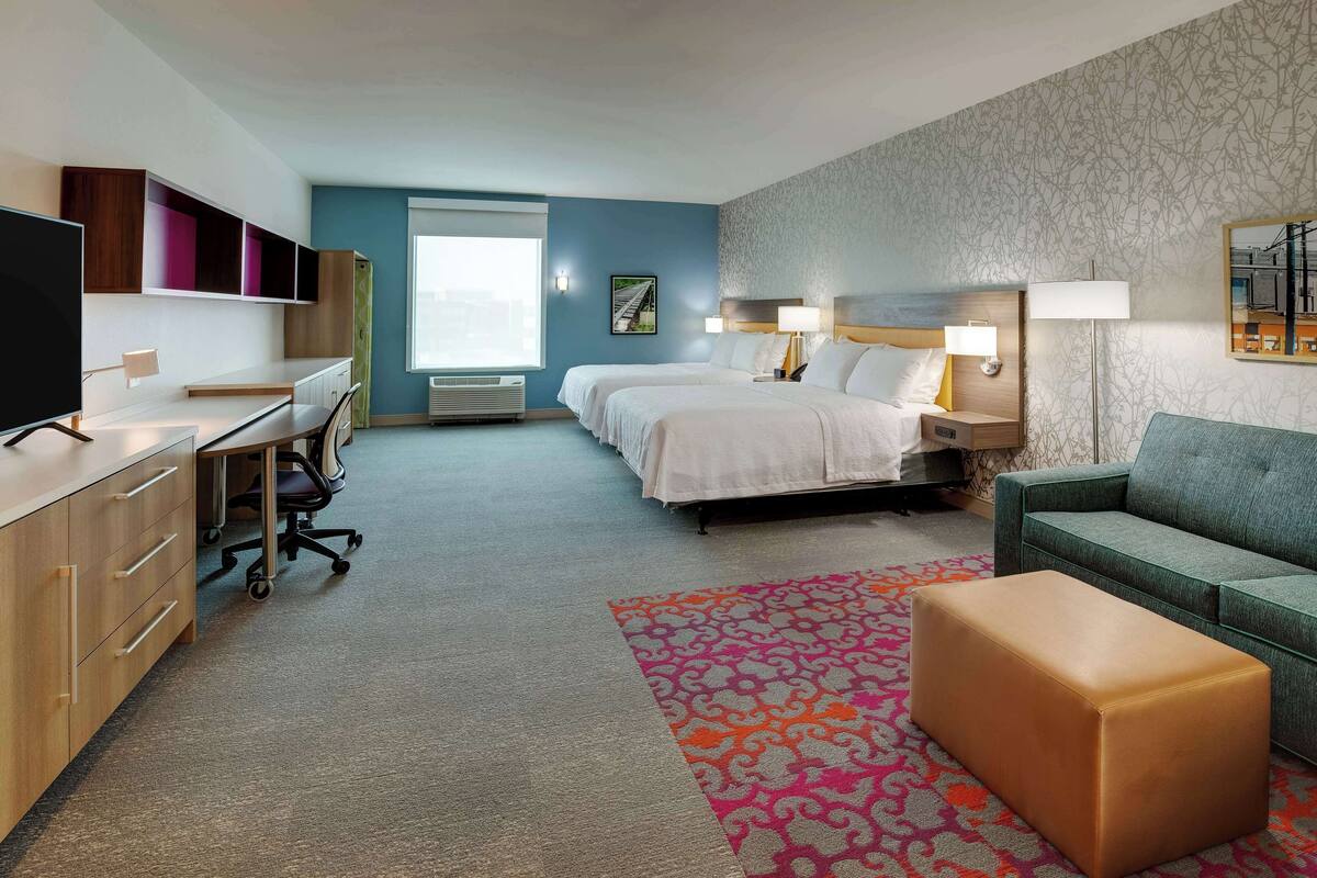 Quarto de hotel com escrivaninha, duas camas, um sofá e um tapete colorido.