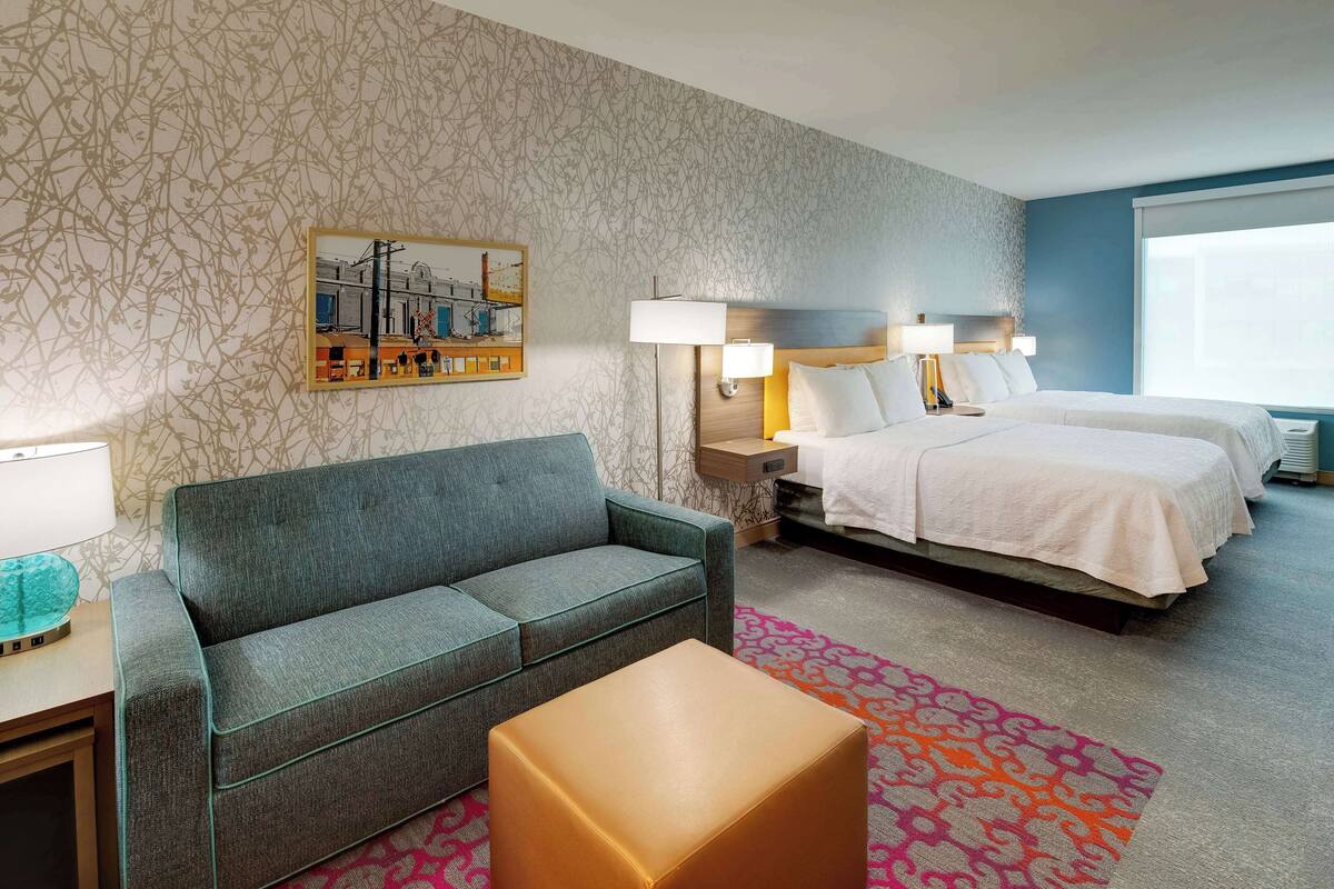 Quarto de hotel com sofá cinzento, cama com lençóis brancos, tapete estampado e um quadro na parede.