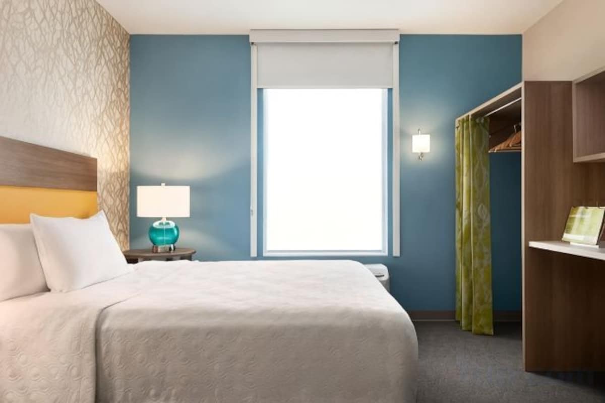 A hotel room with a bed, a nightstand with a lamp, a window with curtains, and a wardrobe.