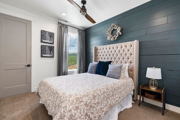 A bedroom with a tufted headboard, a bed with patterned bedding, a nightstand with a lamp, and a view of the outdoors through a window.