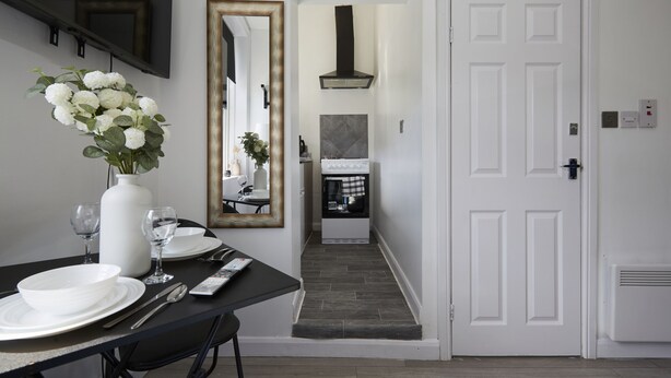 A dining area with a black table set for two, white plates, and a vase of flowers. A large mirror hangs on the wall, and a white door is visible in the background. - Hayy Vibrant Studios Queensgate (Huddersfield)