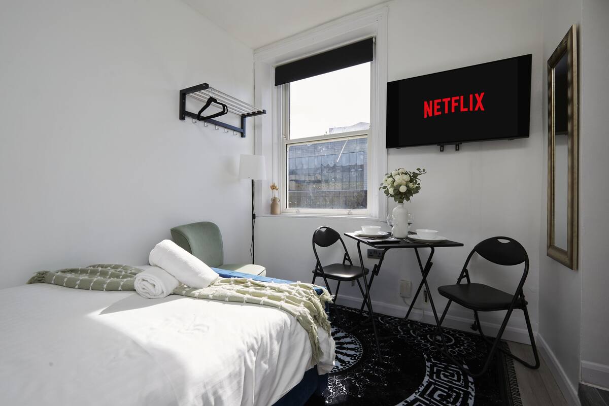 Une chambre d’hôtel comprenant un lit, un bureau avec une chaise, une fenêtre, un téléviseur fixé au mur affichant le logo « NETFLIX », et une petite table sur laquelle est posé un vase de fleurs.