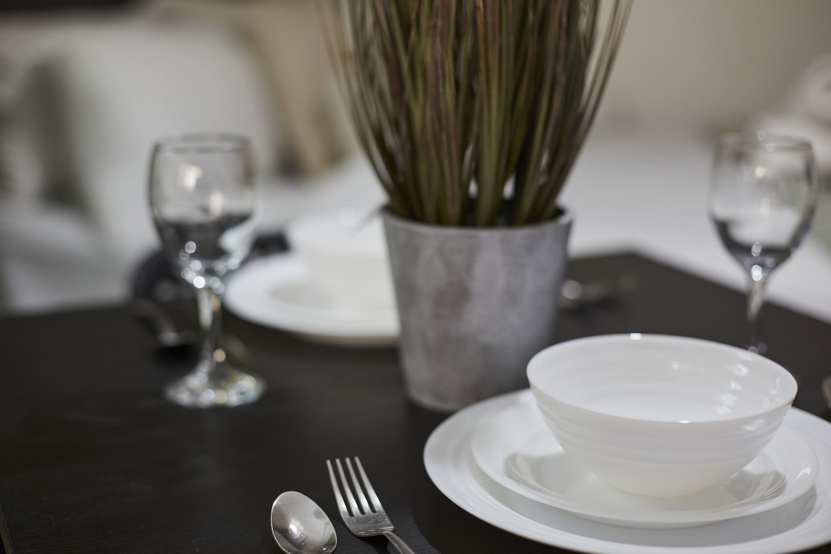 Une table à manger dressée avec des assiettes blanches, un bol, une fourchette, une cuillère et un vase contenant des plantes vertes.
