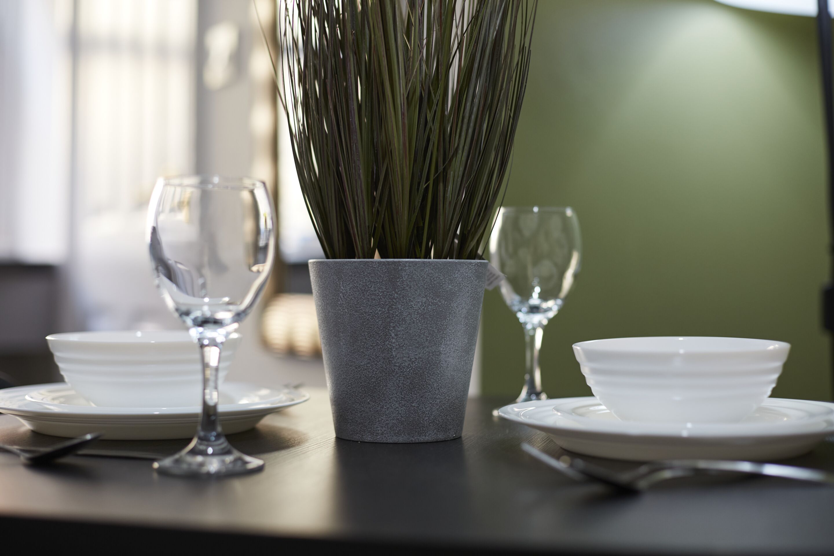 A dining table with a grey pot containing tall green plants, two wine glasses, and white plates.