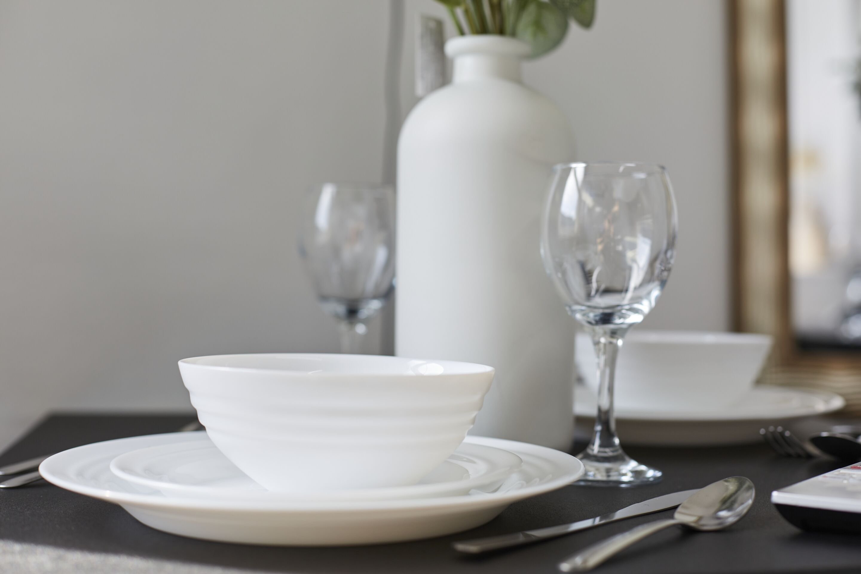 A dining table with a white bowl, a wine glass, and a vase with green plants.