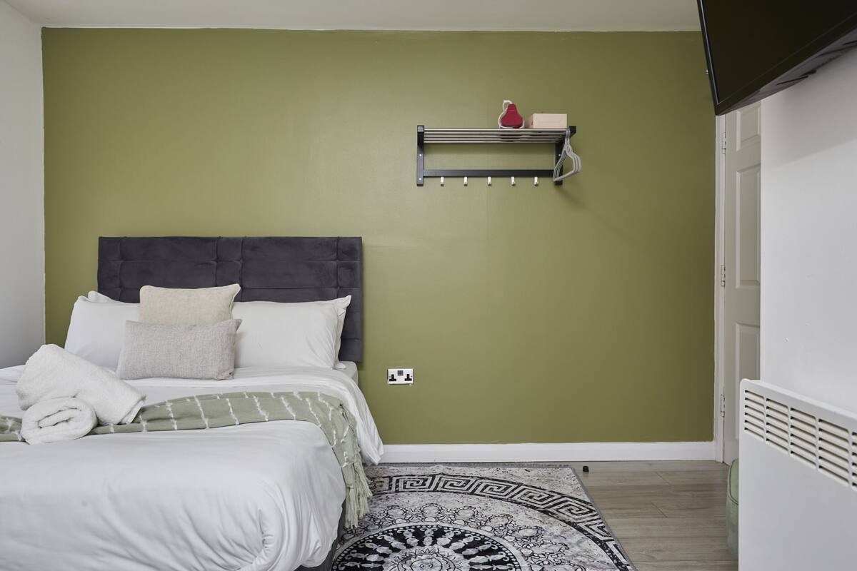 Une chambre à coucher avec un lit, une tête de lit, une étagère fixée au mur sur laquelle se trouve un objet rouge, un tapis à motifs et un radiateur.