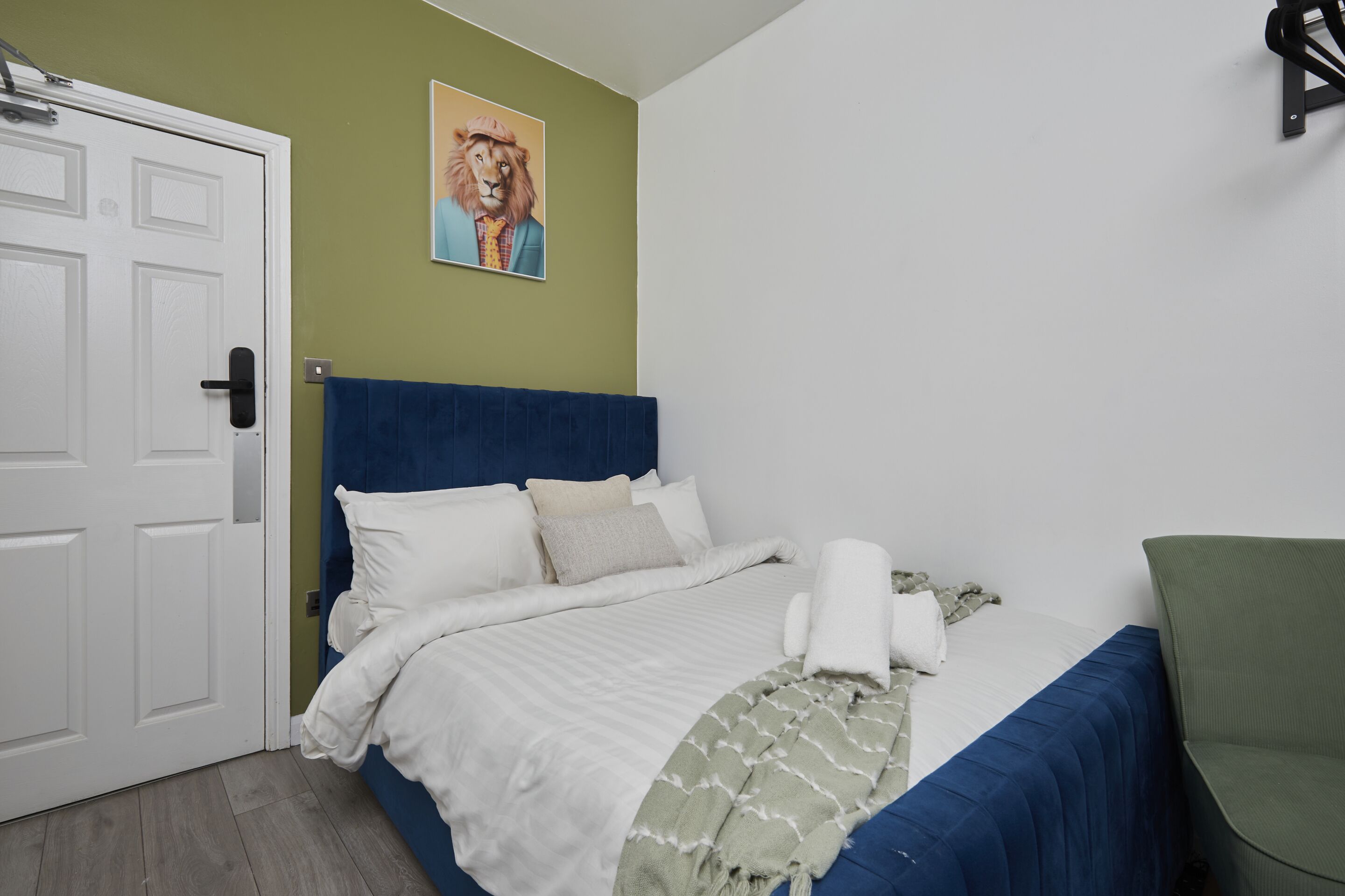 A bedroom with a bed, a green chair, a white door, and a lion painting on the wall.