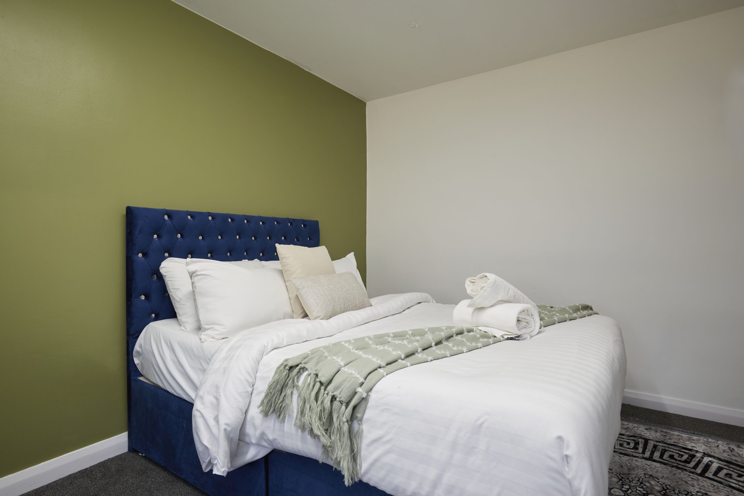 A neatly made bed with white linens, a blue headboard, and a green throw blanket.