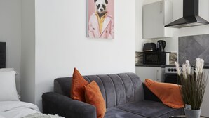A modern living room with a grey sofa, orange cushions, a white bed, a kitchen area with a stove, microwave, and a black range hood. - Hayy Vibrant Studios Queensgate (Huddersfield)