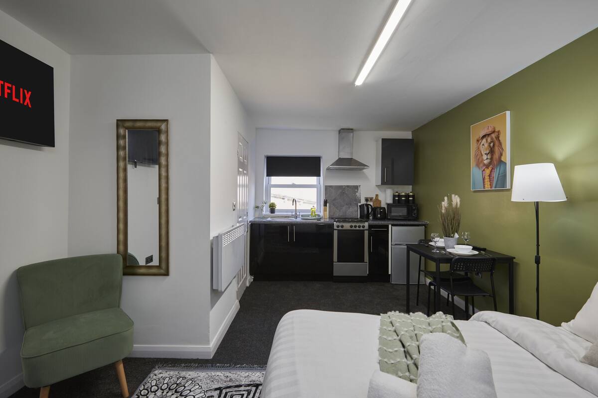 Une chambre d’hôtel moderne comprenant un lit, un fauteuil vert, un petit coin cuisine et une table à manger avec des chaises.