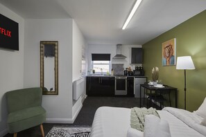 A modern hotel room with a bed, a green armchair, a small kitchen area, and a dining table with chairs. - Hayy Vibrant Studios Queensgate (Huddersfield)