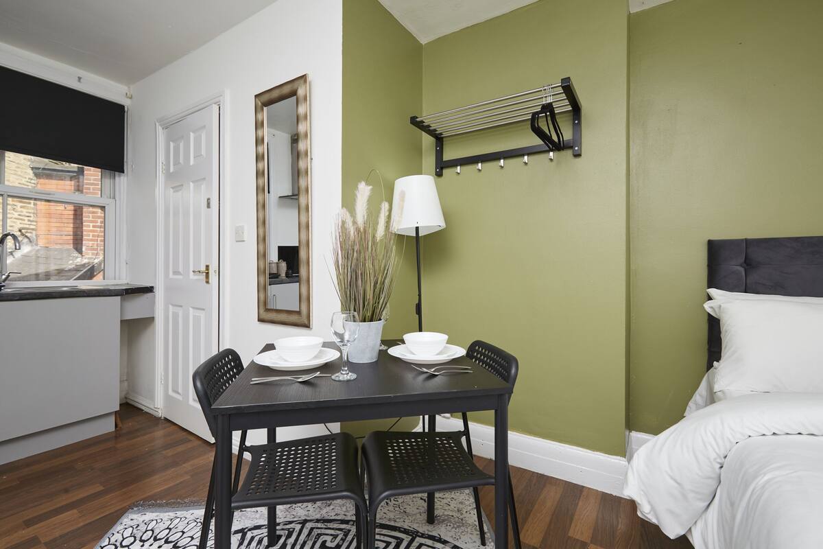Une pièce comprenant une table à manger dressée pour deux personnes, un lit avec une literie blanche, une tête de lit noire, un porte-serviettes fixé au mur et un grand miroir.