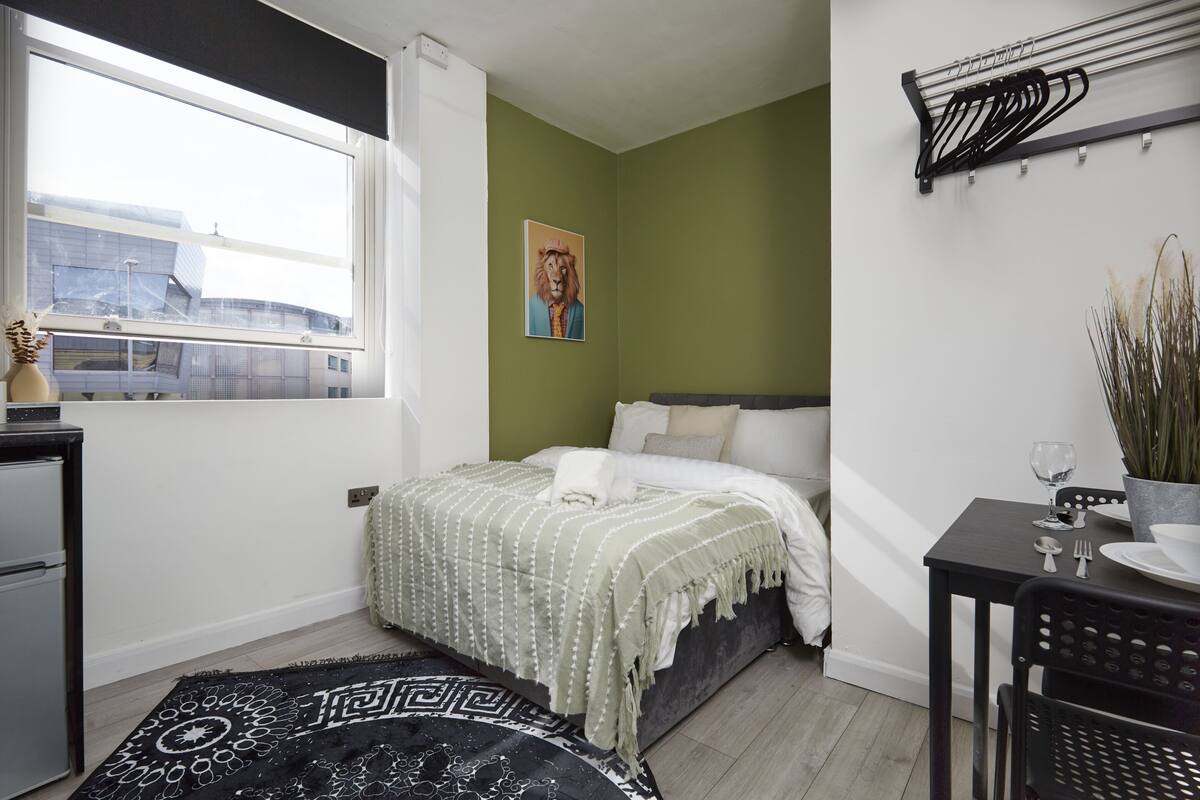 Une chambre à coucher avec un lit, une fenêtre, un porte-manteau fixé au mur et une petite table sur laquelle est posée une plante.