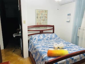 Ein Schlafzimmer mit einem hölzernen Bett, einem gemusterten Bettbezug, zwei aufgerollten Handtüchern und einer wandmontierten Klimaanlage.
