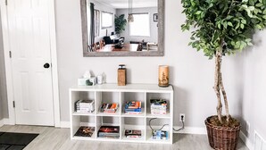 Un pasillo con una puerta blanca, una consola blanca con libros y objetos decorativos, un espejo y una planta en maceta.