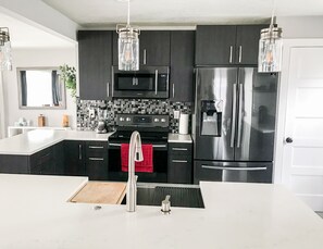 Una cocina moderna con armarios oscuros, electrodomésticos de acero inoxidable, encimera blanca e iluminación colgante.