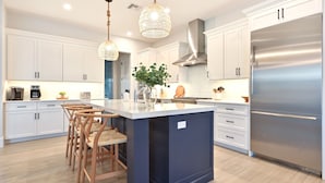 A modern kitchen with white cabinets, a central island with bar stools, stainless steel appliances, and hanging pendant lights. - Hot Tub & Heated Pool, 8 Bikes, 2 King Beds and Suites! (Marco Island)