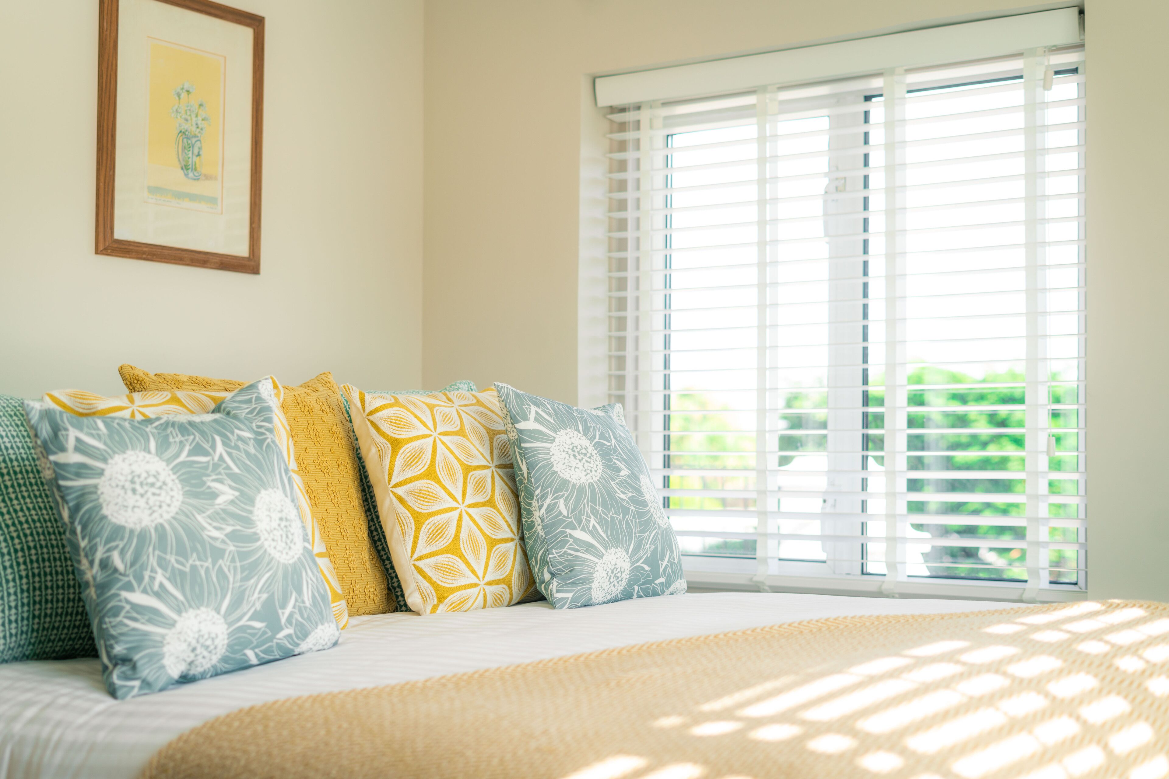 A bedroom with a bed, pillows, a window with blinds, and a framed picture on the wall.