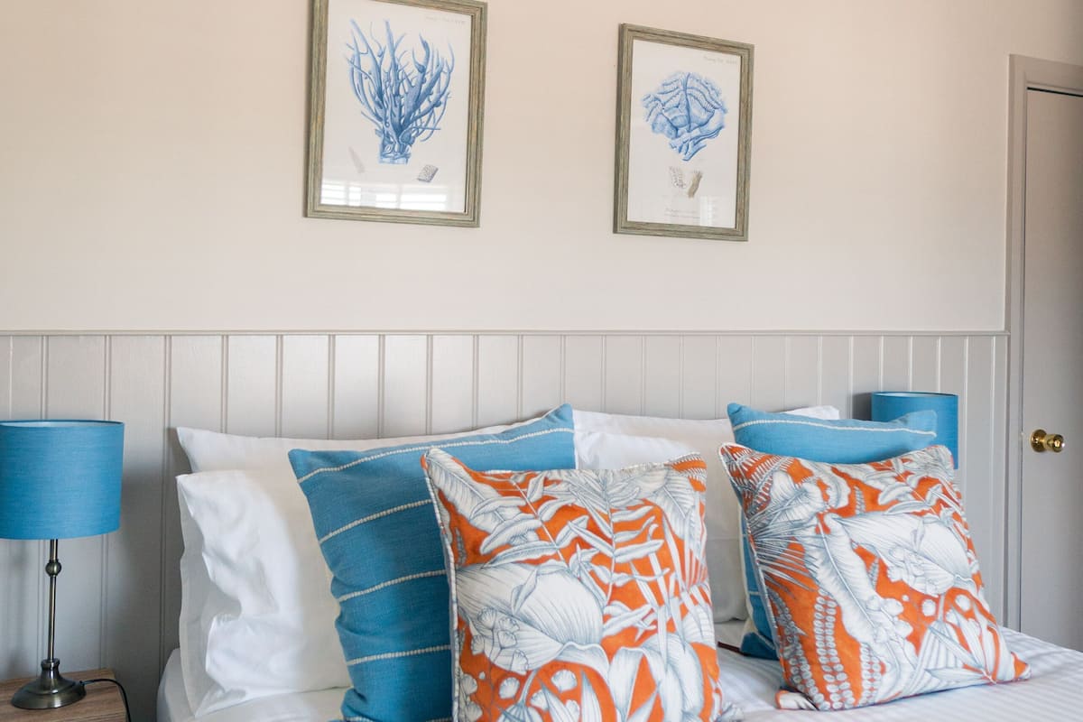 A neatly made bed with a white comforter and blue and orange patterned pillows, a bedside lamp, and two framed wall art pieces.