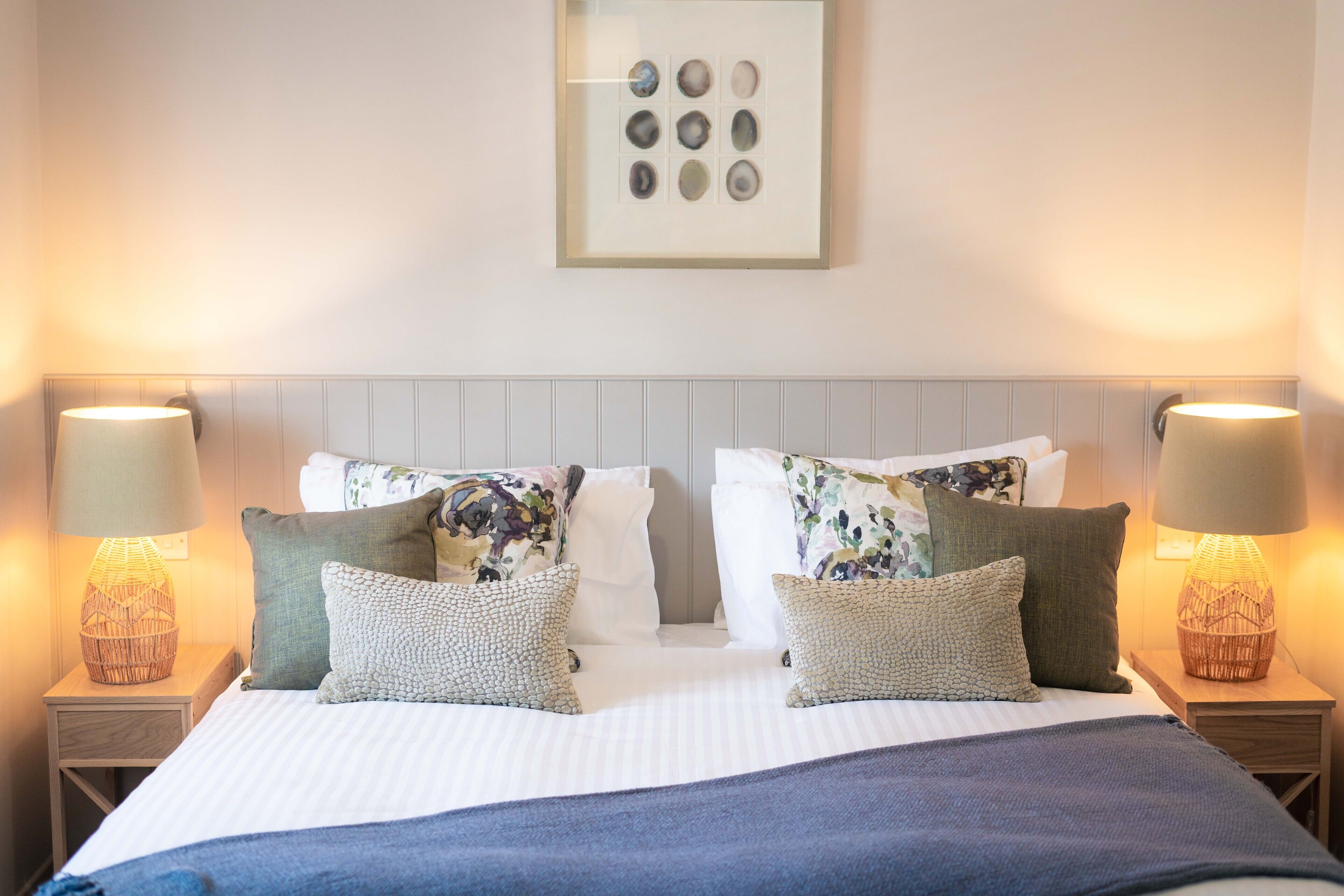 A neatly made bed with floral pillows, a framed artwork above, and two bedside lamps on wooden nightstands.