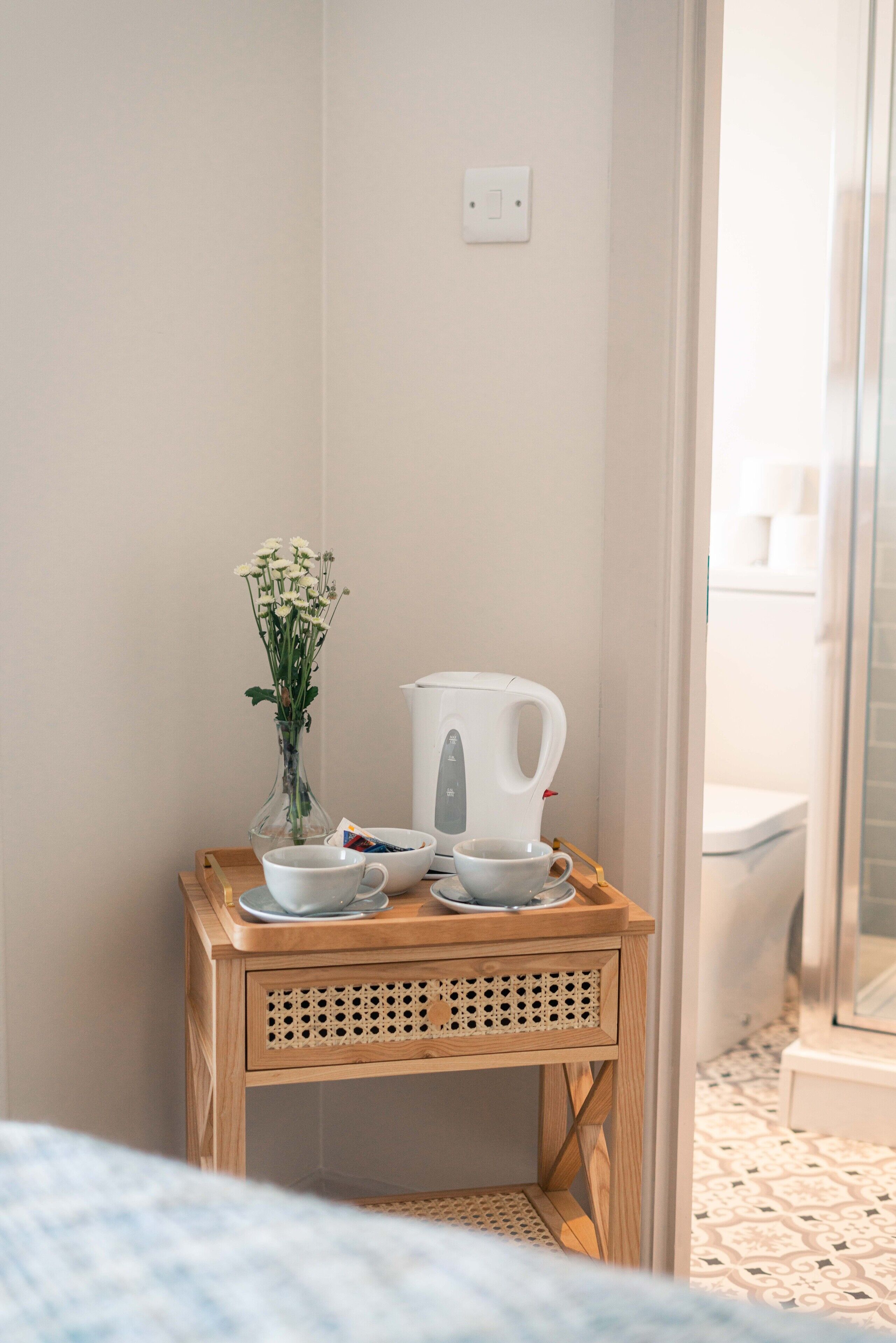 A small wooden table with a white kettle, two cups, and a vase of flowers in a bathroom with a tiled floor and a glass shower door.