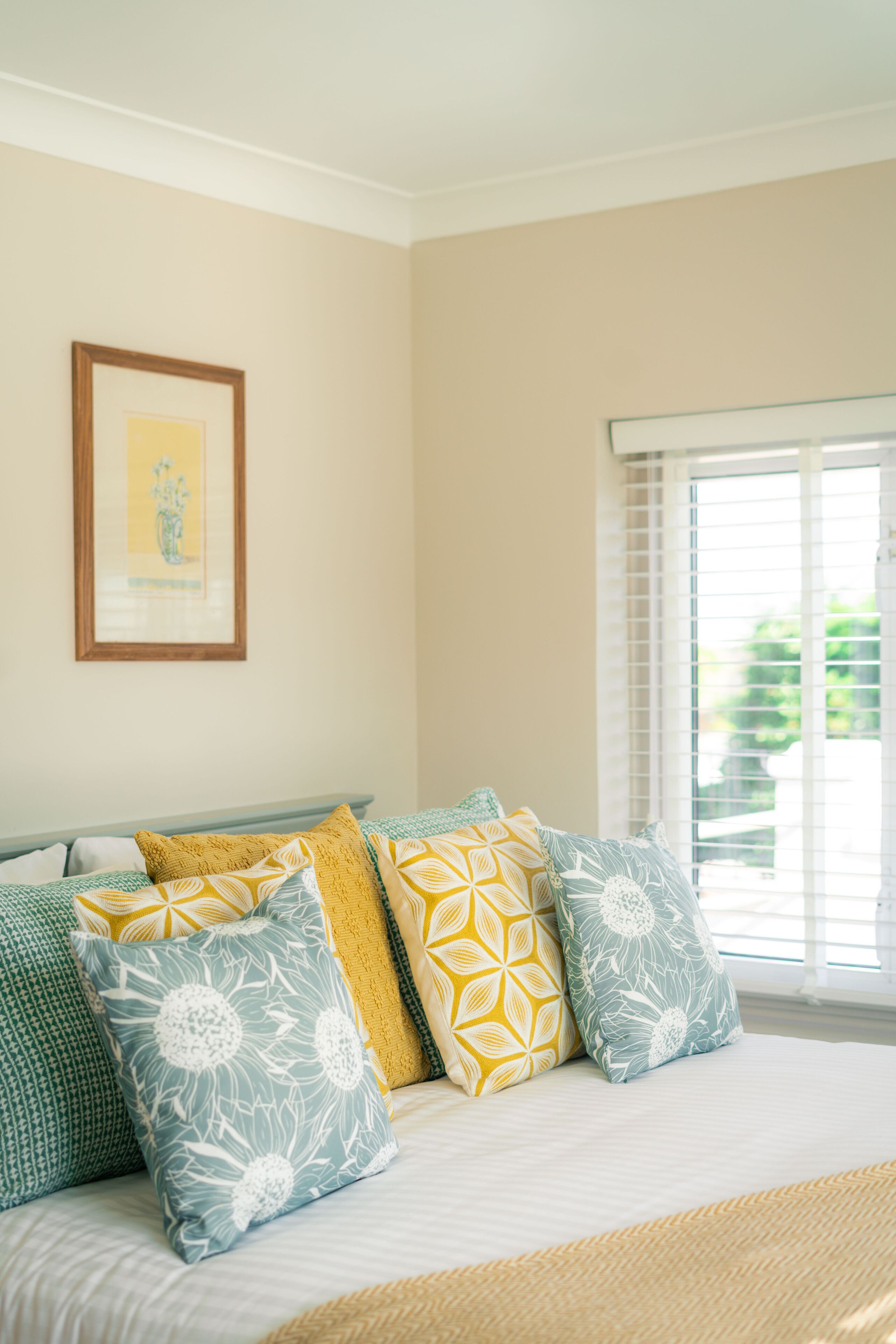 A bedroom with a bed, pillows, a framed picture on the wall, and a window with blinds.