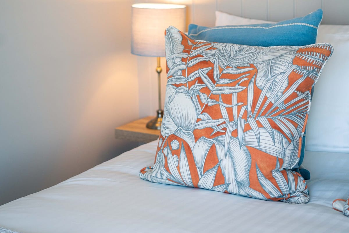 A neatly made bed with white bedding and two pillows, one with a leaf pattern and the other solid blue.