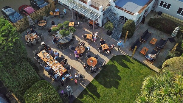 An outdoor dining area with multiple tables and chairs, surrounded by greenery and a building with a sloped roof.