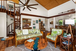 Un salon spacieux doté d’un ventilateur de plafond en bois, d’un escalier en bois, d’un canapé orné d’un motif à feuilles, d’une table basse et d’un coin repas avec des meubles en bois.