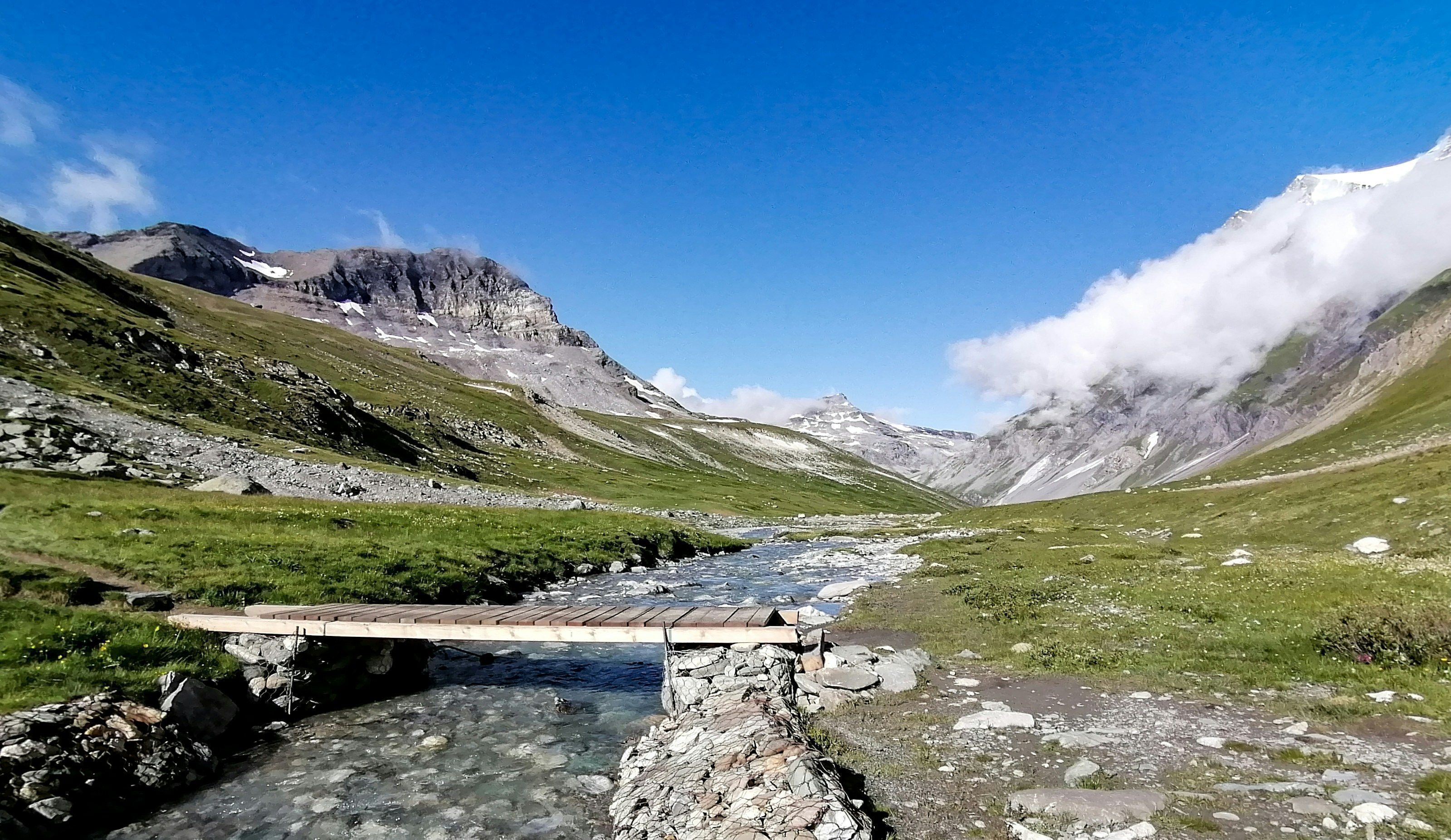 Paesaggio montano con un ponte di legno su un torrente, terreno roccioso e cime innevate.