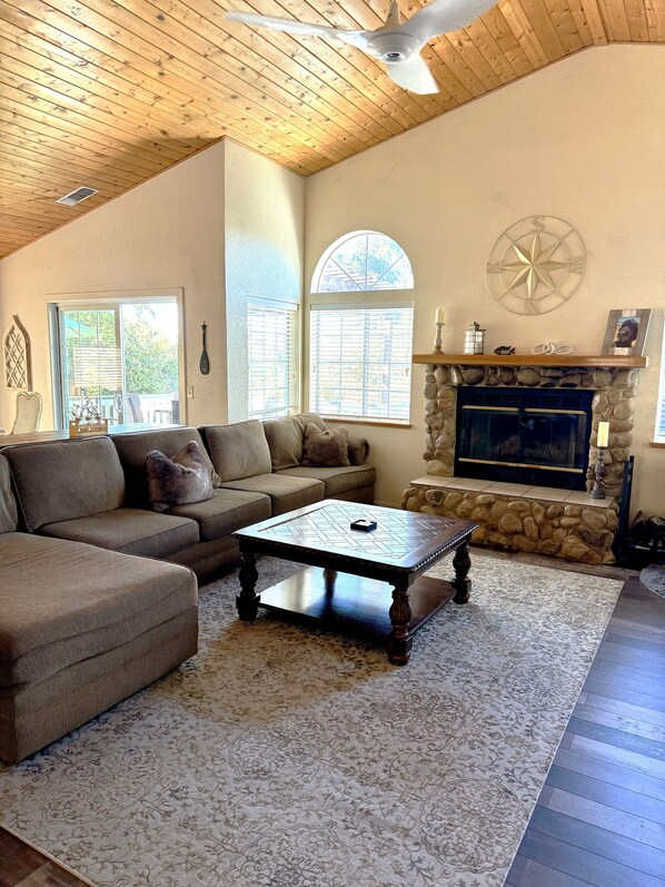 A cozy living room with a stone fireplace, a large window, and a ceiling fan.