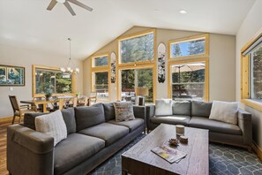 Relax in this spacious living area with a high ceiling, large windows, and a cozy seating arrangement featuring a sectional sofa and a smaller couch.