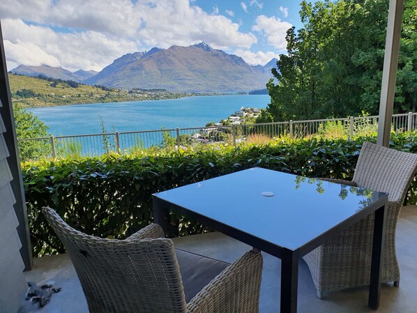 A balcony with wicker chairs and a table overlooking a lake and mountains.