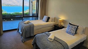 Ein Hotelzimmer mit zwei Betten, einem Nachttisch mit Lampe und einem großen Fenster mit Blick auf das Meer und die Berge.
