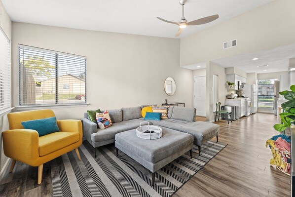 A modern living room with a sofa, ottoman, and yellow armchair. There is a kitchen area visible in the background. - Bright, welcoming home minutes from Q2 + Domain (Austin)