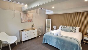 A modern bedroom with a bed, a white chair, a dresser, a mirror, and a wall with vertical wooden slats. - Beautiful Apartment Surrounded by Native Bush (Picton)