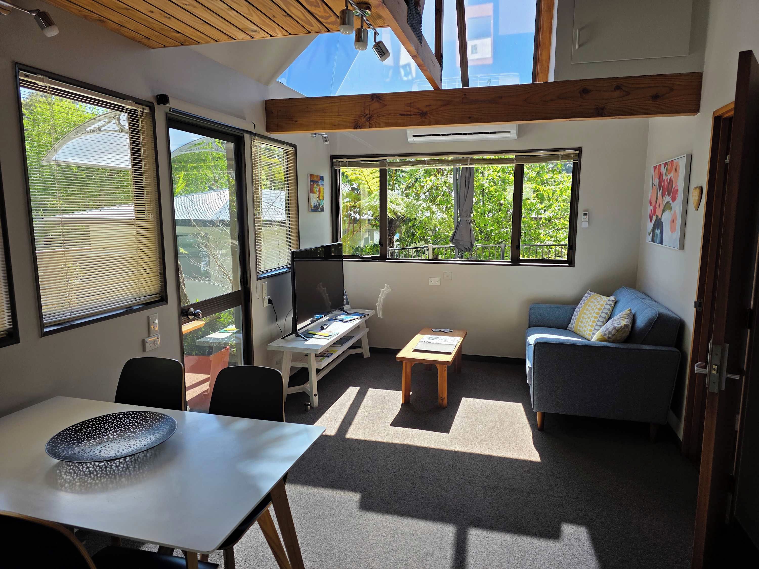 A room with a desk, a chair, a sofa, and a dining table. There are large windows providing natural light and views of greenery outside.