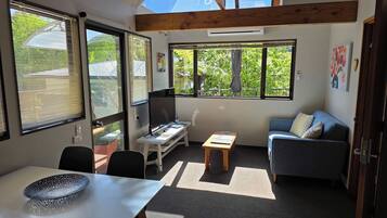 A room with a desk, a chair, a sofa, and a dining table. There are large windows providing natural light and views of greenery outside.