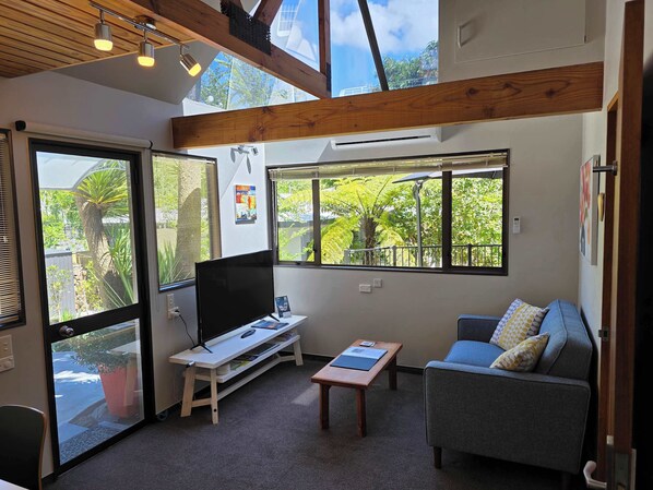 A modern living room with a TV, a sofa, and a coffee table. There are large windows with a view of greenery outside. - Beautiful Apartment Surrounded by Native Bush (Picton)