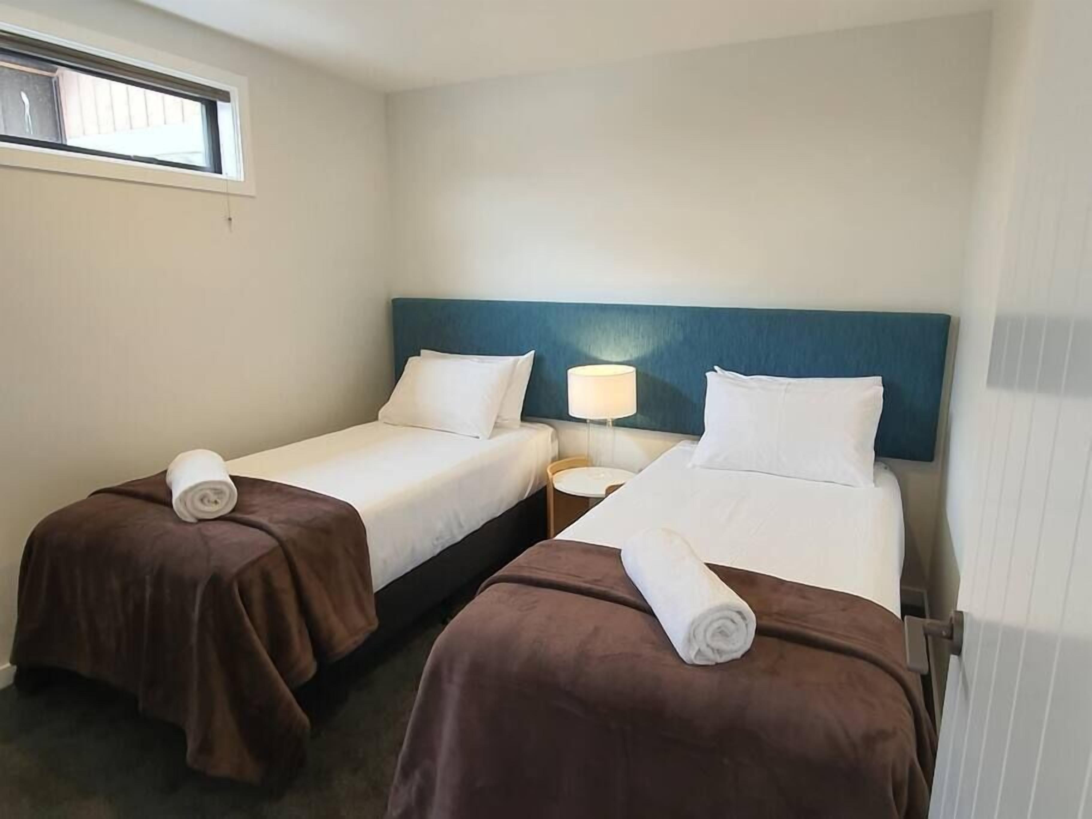 A hotel room with two single beds, each with a brown bedspread and a rolled towel. There is a nightstand with a lamp between the beds.