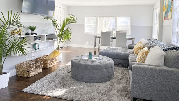A modern living room with a grey sofa set, ottoman, and a flat-screen TV mounted on the wall. There are two potted plants, a wicker basket, and a fluffy rug on the wooden floor.