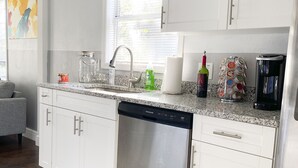 A modern kitchen with white cabinets, a stainless steel dishwasher, a granite countertop, and a window with blinds.