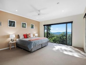 A bedroom with a bed, two bedside tables, a ceiling fan, and a large window with a view of greenery.