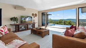 A modern living room with a sofa, coffee table, TV, and a large sliding glass door leading to a balcony with a view. - Wilderness House l Infinity Pool & Views (Kulangoor)