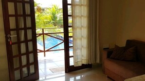 A room with a sofa, a wooden door with glass panels, and a view of a swimming pool and palm trees outside. - Hospedagens La Fazenda Canoa Quebrada (Aracati)