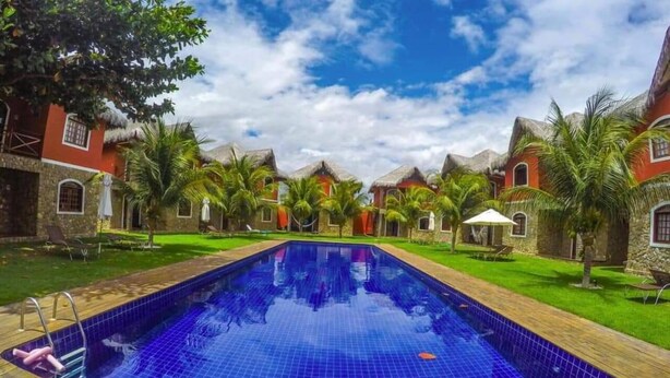 A swimming pool with a wooden deck, surrounded by palm trees and buildings with red and stone facades. - Hospedagens La Fazenda Canoa Quebrada (Aracati)