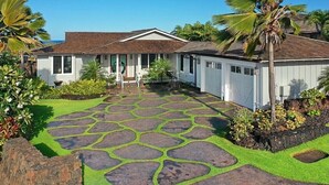 A house with a stone patio, a white exterior, and a well-maintained garden with palm trees. - Gorgeous Poipu Villa with Pool & Spa + Ocean Views (Koloa)